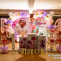 A fairy and butterfly-themed first birthday with a personalized backdrop featuring the child's photo. The decor includes themed plinths with mushroom and fairy graphics, a light-up number '1', and a pink and purple balloon arch.