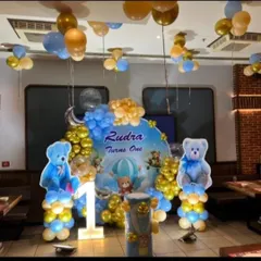 A sweet and cuddly teddy bear theme for a first birthday. This decoration features a "Rudra Turns One" backdrop, large bear cutouts, a light-up number '1', and a blue and gold balloon arch.