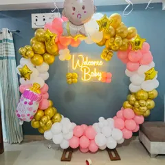 This is a modern and stylish setup for a baby shower. It features a circular balloon wreath in pink, white, and gold, with a 'Welcome Baby' neon sign that looks great in photos.