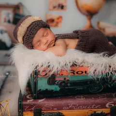 A beautiful side profile of the sleeping newborn, resting on a soft fur blanket atop the suitcases. The knitted outfit and hat add a cozy, handmade touch to the portrait.