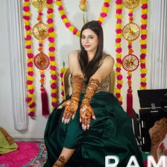 A happy client posing with her completed bridal henna in front of a colorful mehndi ceremony backdrop.