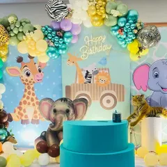 A colorful and kid-friendly jungle theme birthday party. The backdrop features cute cartoon animals like a giraffe and elephant, surrounded by a vibrant balloon arch with animal print balloon accents.