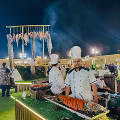 Our impressive live seafood station, where guests can see the fresh catch of the day. Here, a chef proudly presents a large grilled fish, with a backdrop of hanging fish and the sizzle of the grill, promising a fresh coastal feast.
