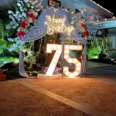 A grand celebration for a 75th birthday. This outdoor setup features large, brightly lit marquee numbers, a 'Happy Birthday' neon sign, and an arch decorated with white drapes and flowers for an elegant touch.