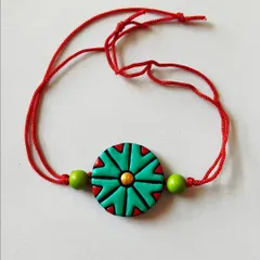 A terracotta rakhi with a geometric green and red design. These rakhis are handmade and painted, making each one a special token of love.