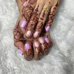 A lovely gel polish application on natural nails. This design features a soft lavender color on some nails and a nude base with a hint of glitter on others, perfect for a subtle look against mehndi.