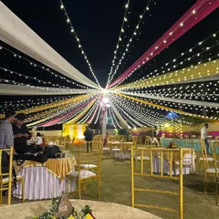 A spectacular canopy of colorful drapes and fairy lights for an outdoor Haldi event. The vibrant yellow, pink, and white fabrics radiate from a central point, creating a festive tent-like feel over the entire lawn.