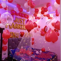 A romantic bedroom decorated in red and white for a newly married couple. The room features "Just Married" foil balloons, heart-shaped balloons, and a delicate canopy over the bed illuminated with warm fairy lights.