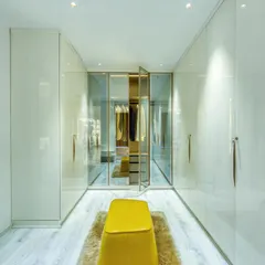 A spacious walk-in closet with sleek, handleless wardrobes and a central yellow ottoman on a plush rug. The glass-doored section allows for curated display.