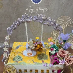 This is my signature Teej Hamper, a symbol of love, tradition, and festivity. It features a beautiful swing with miniature dolls, a box of traditional Ghewar, and other festive essentials, all presented in an elegant basket to make the celebration truly magical.