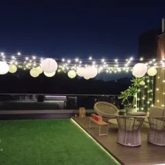 A wide view of a terrace decorated for an evening party. The space is illuminated by a canopy of string lights and hanging paper lanterns, creating a warm and inviting ambiance over the modern outdoor seating area.