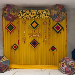 This is a classic Haldi backdrop with a yellow curtain, a "Haldi" sign, and colorful Rajasthani umbrellas. The setup is bright and perfect for capturing the joyful moments of the ceremony.