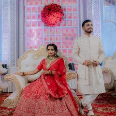 A regal portrait of the couple at their reception. The bride is seated on a throne-like chair while the groom stands beside her, creating a classic and elegant wedding portrait.