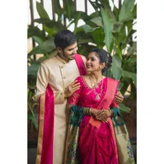 A classic couple portrait showcasing beautiful Maharashtrian wedding attire. The vibrant pink and gold of their outfits create a stunning visual against the natural green background.