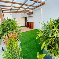 The completed terrace garden, now a vibrant social space. It features a glass-roofed pergola, a custom bar area, and lush greenery in planters, all framed by artificial turf for a low-maintenance, always-green look.