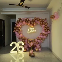 A stylish 26th birthday setup featuring a heart-shaped balloon arch in rose gold and pink. The look is completed with a "Happy Birthday" neon sign and large marquee numbers.