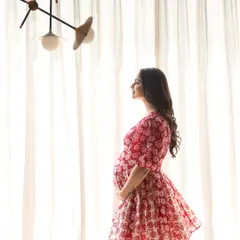 Bathed in the soft light of a window, the mother-to-be stands in profile, her red and white dress adding a pop of color to this serene, at-home portrait.