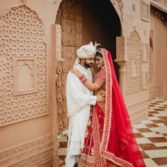 An intimate embrace against a stunning architectural backdrop. The groom holds his bride close, her red lehenga a beautiful pop of color against his white sherwani, creating a portrait of royal elegance.
