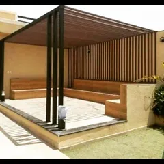 This pergola features a dark metal frame with a slatted roof and back wall, paired with built-in wooden bench seating. The design creates a defined, shaded seating area within a larger garden.