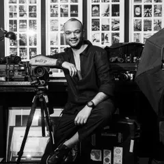 A black and white portrait of photographer Pushkar Thakur in his studio. His relaxed smile and the backdrop of his own work and equipment give a real sense of him as a creator.