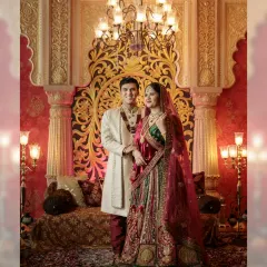 The couple poses against a lavishly decorated backdrop with rich red and gold details. Their traditional outfits and the opulent setting evoke the grandeur of Indian royalty.
