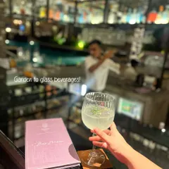 A guest holds a freshly made cocktail at the Asri bar counter. This image captures the "garden-to-glass" experience, with the bartender visible in the background, shaking up another creation.