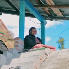 An elderly woman sits peacefully on the steps of her porch in a Himachali village. The simple, rustic architecture and the clear blue sky in the background frame a portrait of contentment and a life connected to the mountains.