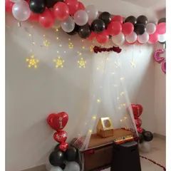 A lovely canopy setup perfect for an anniversary or romantic surprise. The decor includes a balloon garland on the ceiling, star-shaped lights, and a trail of rose petals on the floor.