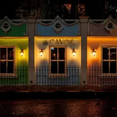 The exterior facade of Los Cavos at night, designed to look like a row of colourful colonial buildings. Each section is painted a different hue and lit by traditional wall sconces, creating a vibrant and welcoming entrance that sets the tone for the experience inside.