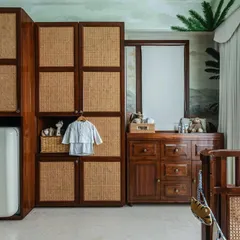 Functionality is key in a nursery. Here you can see the combination of rattan and wood cabinetry, providing ample storage, alongside a classic wooden changing table and a retro-style Smeg fridge for convenience.