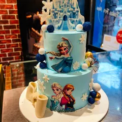 A two-tier Frozen cake with an edible ice castle topper. This design captures the magic of the movie with characters Elsa, Anna, and Olaf, decorated with shimmering blue and white spheres.