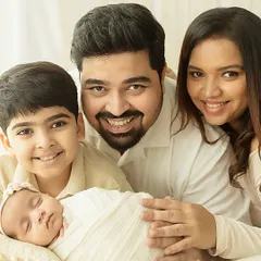A classic family-of-four pose, with everyone smiling at the camera. The sleeping newborn adds a touch of serenity to this happy family portrait, captured in a bright, welcoming studio setting.