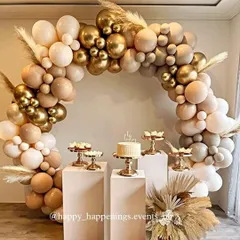 A stunning boho-themed balloon arch in neutral and gold tones. This setup, complete with dessert plinths and pampas grass accents, is perfect for creating a chic and trendy atmosphere for any celebration.