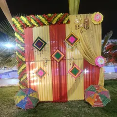 To create a traditional Rajasthani feel, I use elements like these colorful folk art hangings and decorative umbrellas. The striped yellow and red fabric backdrop makes the whole setup feel very festive.