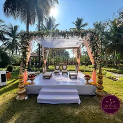 An elegant outdoor 'Muhurtham Mantap' set against a beautiful backdrop of palm trees. The simple white stage, pastel drapes, and fresh floral arrangements create a serene and picturesque setting for the wedding ceremony.