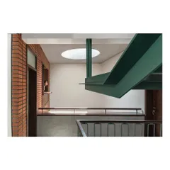 The main staircase is a central sculptural element within the Nairy Residence. A bold, green-painted steel beam supports the structure, creating a visual contrast with the raw brick wall and drawing the eye upwards towards a circular skylight, which bathes the double-height volume in daylight.