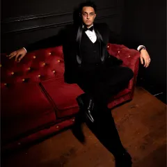 A dapper groom's portrait. The dark, moody lighting and the confident pose on the red sofa create a very stylish, editorial feel.