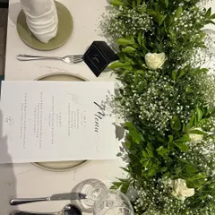 An overhead view of a complete place setting. The custom menu, crisp white napkin, and polished silverware are beautifully arranged alongside the fresh greenery of the table runner for a clean and sophisticated look.