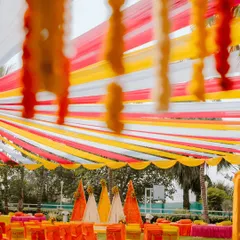 A wide view of a vibrant Haldi ceremony we designed. The space is brought to life with a canopy of colourful drapes in yellow, red, and white, complemented by marigold flower strings and cone-shaped floral backdrops, creating a festive and energetic outdoor setting.