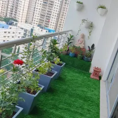 A full view of a balcony transformation we completed. It features a row of flowering rose plants in grey planters, a serene Buddha statue, and a lush artificial grass floor.