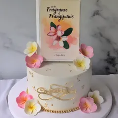 A beautiful and elegant cake designed for a book launch. The centerpiece is an edible replica of the book cover for "In the Fragrance of Frangipanis," surrounded by handcrafted sugar flowers that match the theme.