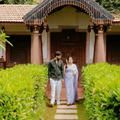 A couple dressed in beautiful ethnic wear, walking hand-in-hand down a path towards a traditional Indian-style home.