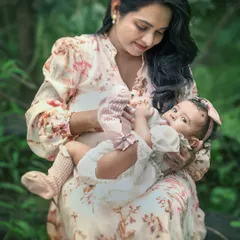 A mother gazes lovingly at her baby girl resting in her lap during an outdoor photoshoot. The soft lighting and lush green surroundings create a peaceful and intimate portrait of motherhood.