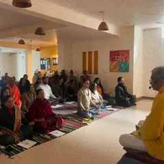 I guide a large group through meditation during a special satsang. My role is to hold the space and offer guidance, allowing each person to connect with their own inner stillness.