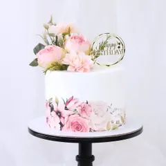 A pretty and elegant birthday cake featuring a printed floral wrap on the bottom tier. The design is topped with a delicate arrangement of fresh pink flowers and a "Happy Birthday" topper.