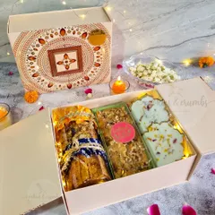 An inside look at one of my premium Diwali hampers. This beautiful box contains a jar of assorted nuts and treats, a walnut tea cake loaf, and flower-shaped white chocolates, all nestled with festive fairy lights.