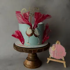 A unique celebration cake with hand-painted wine glasses clinking in a toast. The design is set against a cool blue background and accented with abstract, vibrant red sugar sails for a modern, artistic flair.
