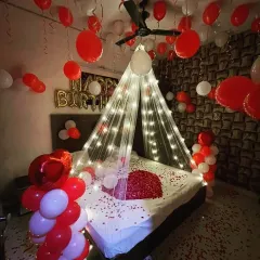 A cozy and romantic birthday setup with a lighted canopy over the bed, red and white balloons, and scattered rose petals.