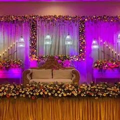 An elegant wedding stage design with a beautiful floral frame and dramatic purple uplighting. The combination of hanging birdcage lamps, candle stands, and a plush sofa creates a sophisticated and grand backdrop for the couple.