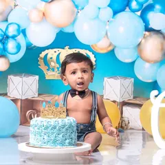 A little prince celebrating his first birthday with a royal blue cake smash. The setup includes a crown, balloons, and a delicious-looking cake ready to be smashed.